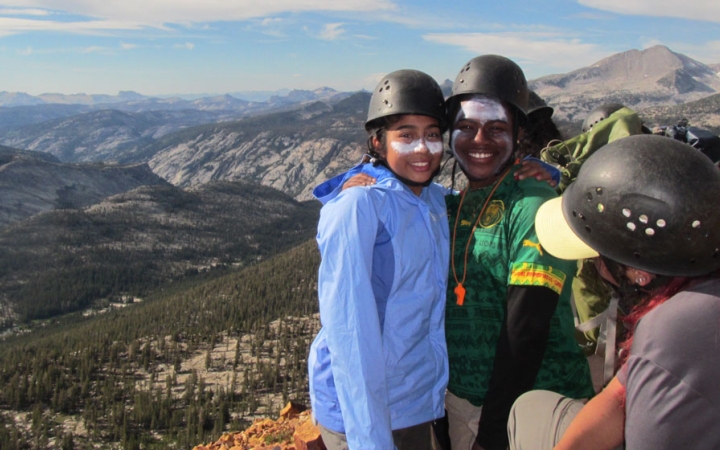 two people wearing helmets and sunscreen smile for a photo with a mountainous landscape behind them